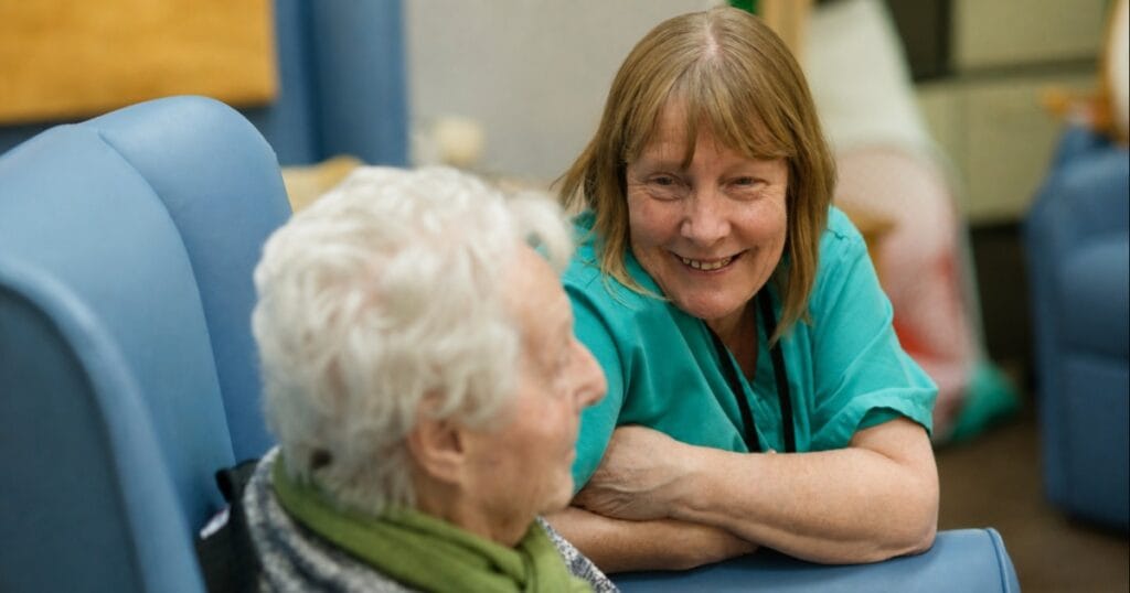 Two women smiling and talking while seated in blue chairs in a care home setting