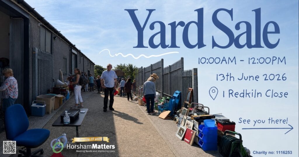 Yard sale sign with people browsing tables and items along a housing estate path on a sunny day