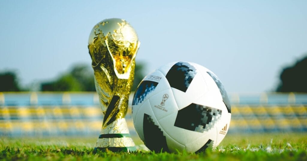 A football next to a golden trophy on a grassy pitch with stadium seats in the background