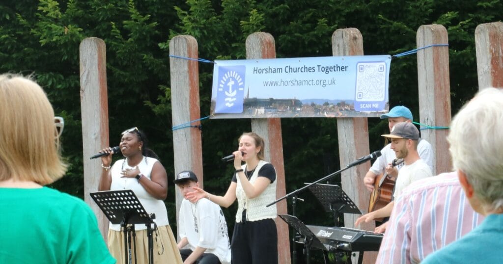 Outdoor church performance with a woman singing into a microphone, a man speaking beside her, musicians in the background, and people watching in front of a banner with church text