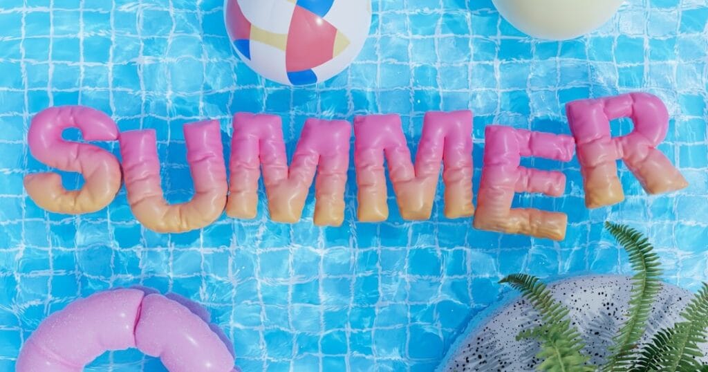Summer letters floating in a swimming pool with a beach ball and inflatable ring alongside palm leaves