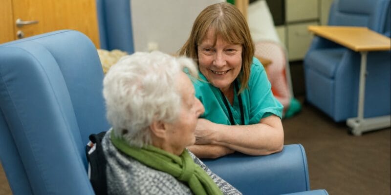 Woman smiling and talking with an elderly woman in a care home lounge