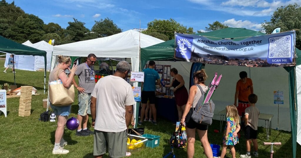 People standing around white and green gazebos at an outdoor community stall on a sunny day, with children and balloons in the foreground