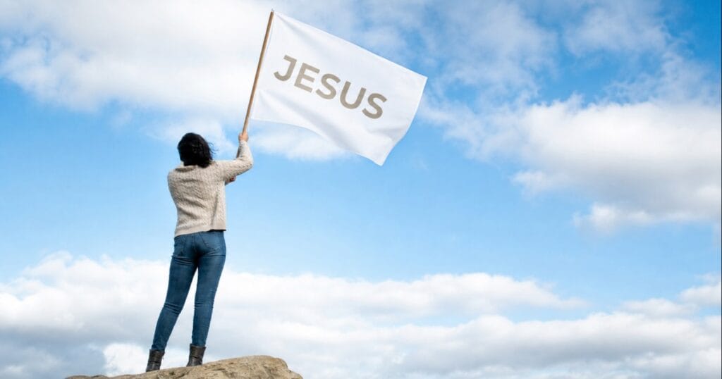 A person on a rocky hill holding up a white flag with the word Jesus against a blue sky