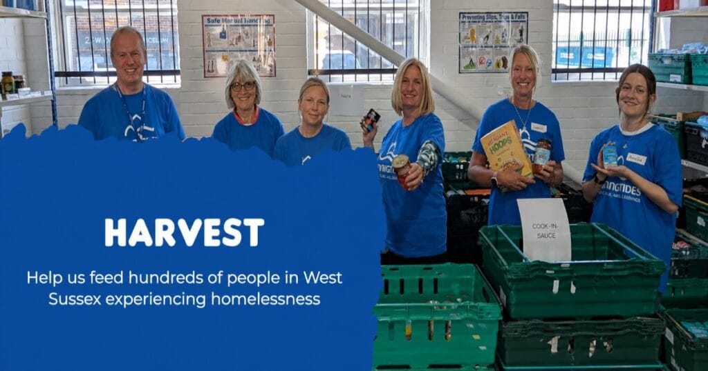 Six people in blue shirts standing behind stacks of green food crates in a warehouse, holding food items, with a Harvest banner in the background