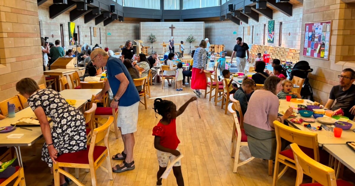 People and children gathered around tables in a bright hall with a cross at the front