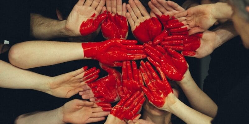 Multiple hands with red paint forming a circle against a dark background