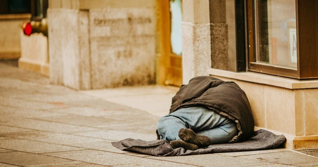 Person sleeping on a sidewalk beside a building, covered with a dark blanket