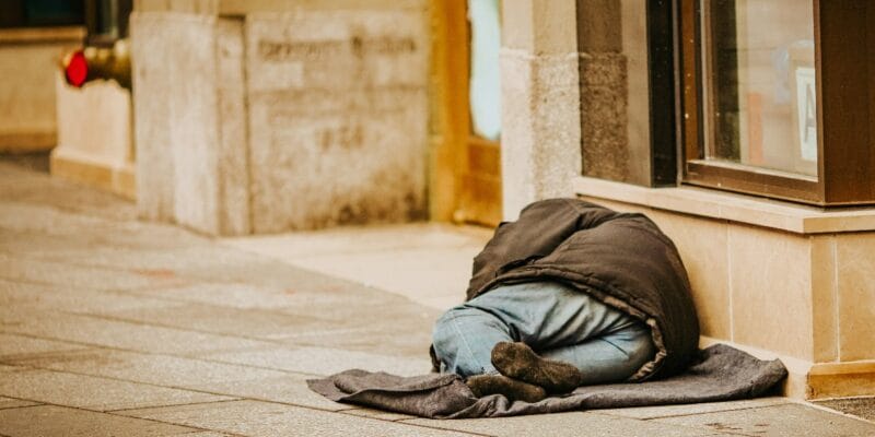 Person sleeping on a sidewalk beside a building, covered with a dark blanket