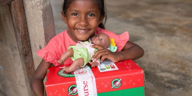 Smiling child holding a doll on a red and green cardboard box
