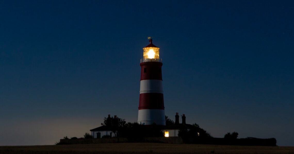Red-and-white striped lighthouse with its lamp illuminated at dusk